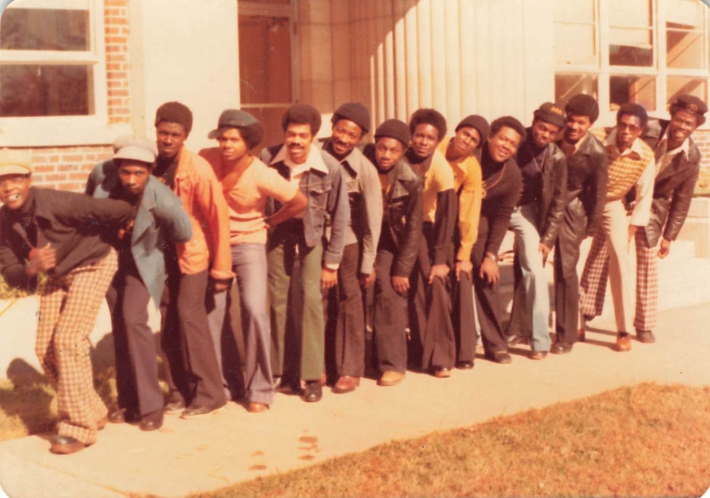 A line of brothers in 1970s dress posed in a row outside a campus building