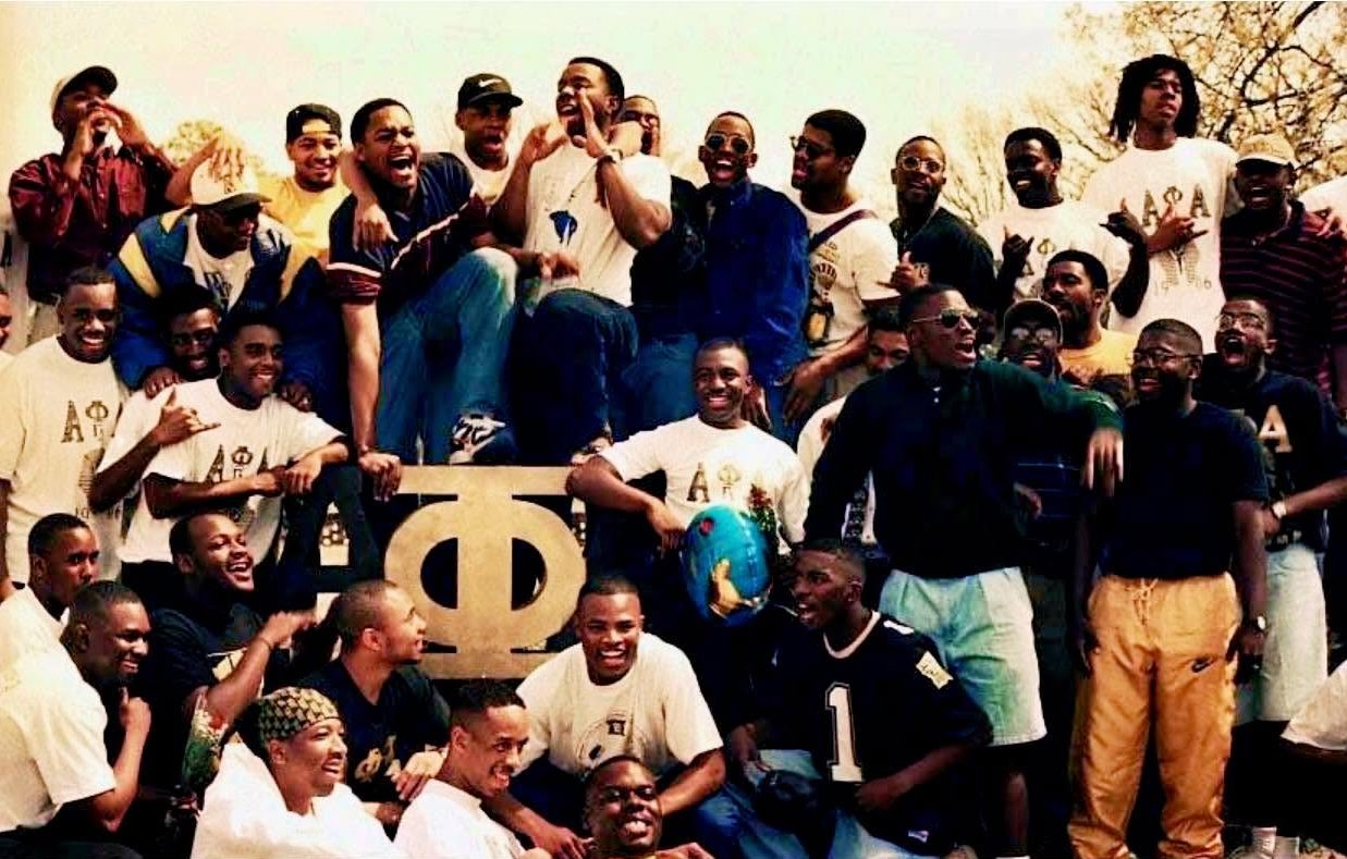 Brothers in Alpha Phi Alpha shirts cheering around a large gold Phi, Spring 1995
