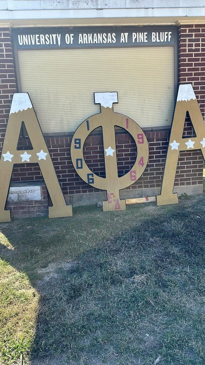 Gold Alpha Phi Alpha letters marked 1906 and 1946 on the lawn below a University of Arkansas at Pine Bluff sign